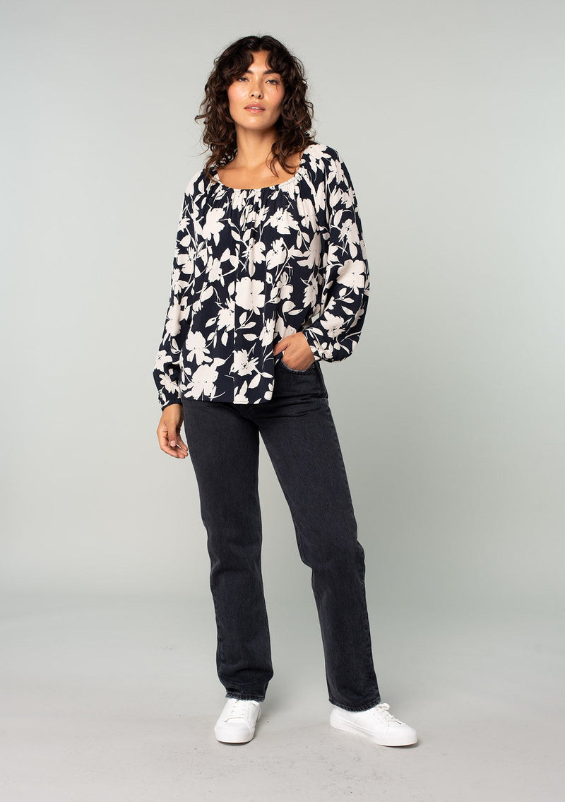 [Color: Black/Natural] A full body front facing image of a brunette model wearing a black and off white floral print blouse. A bohemian resort top with long sleeves and a wide elastic neckline. 