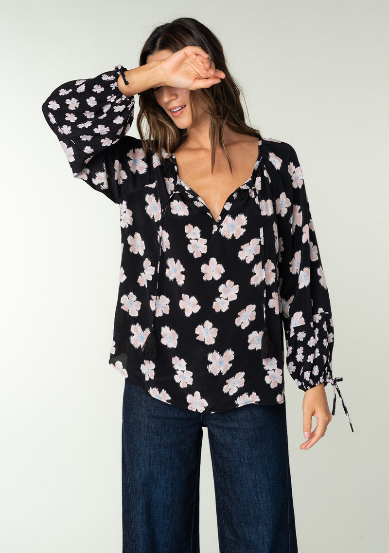 [Color: Black/Taupe] A half body front facing image of a brunette model wearing a flowy bohemian top in a black and taupe floral print. With long sleeves, adjustable tie wrist cuffs, and a split v neckline with tassel ties. 