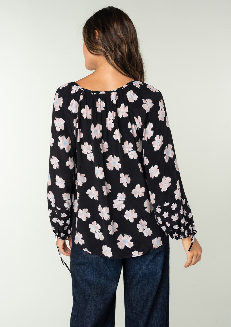 [Color: Black/Taupe] A back facing image of a brunette model wearing a flowy bohemian top in a black and taupe floral print. With long sleeves, adjustable tie wrist cuffs, and a split v neckline with tassel ties. 