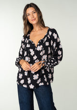 [Color: Black/Taupe] A front facing image of a brunette model wearing a flowy bohemian top in a black and taupe floral print. With long sleeves, adjustable tie wrist cuffs, and a split v neckline with tassel ties. 