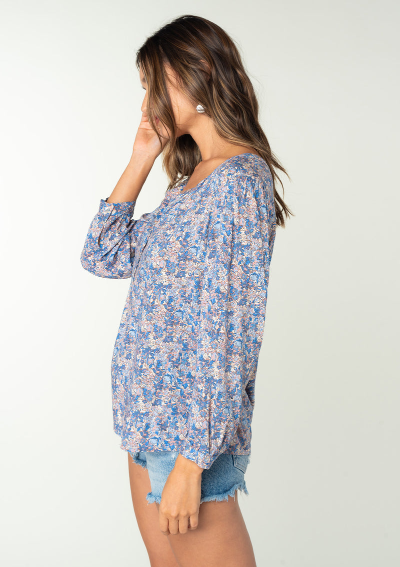 [Color: Blue/Coral] A side facing image of a brunette model wearing a blue and coral floral print bohemian blouse. With long sleeves and a button front. 