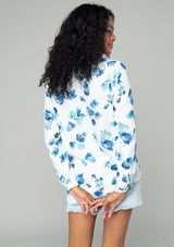 [Color: Ivory/Aqua] A back facing image of a brunette model wearing a classic long sleeve blouse in an ivory white and blue floral print. With a v neckline and button wrist cuff. 