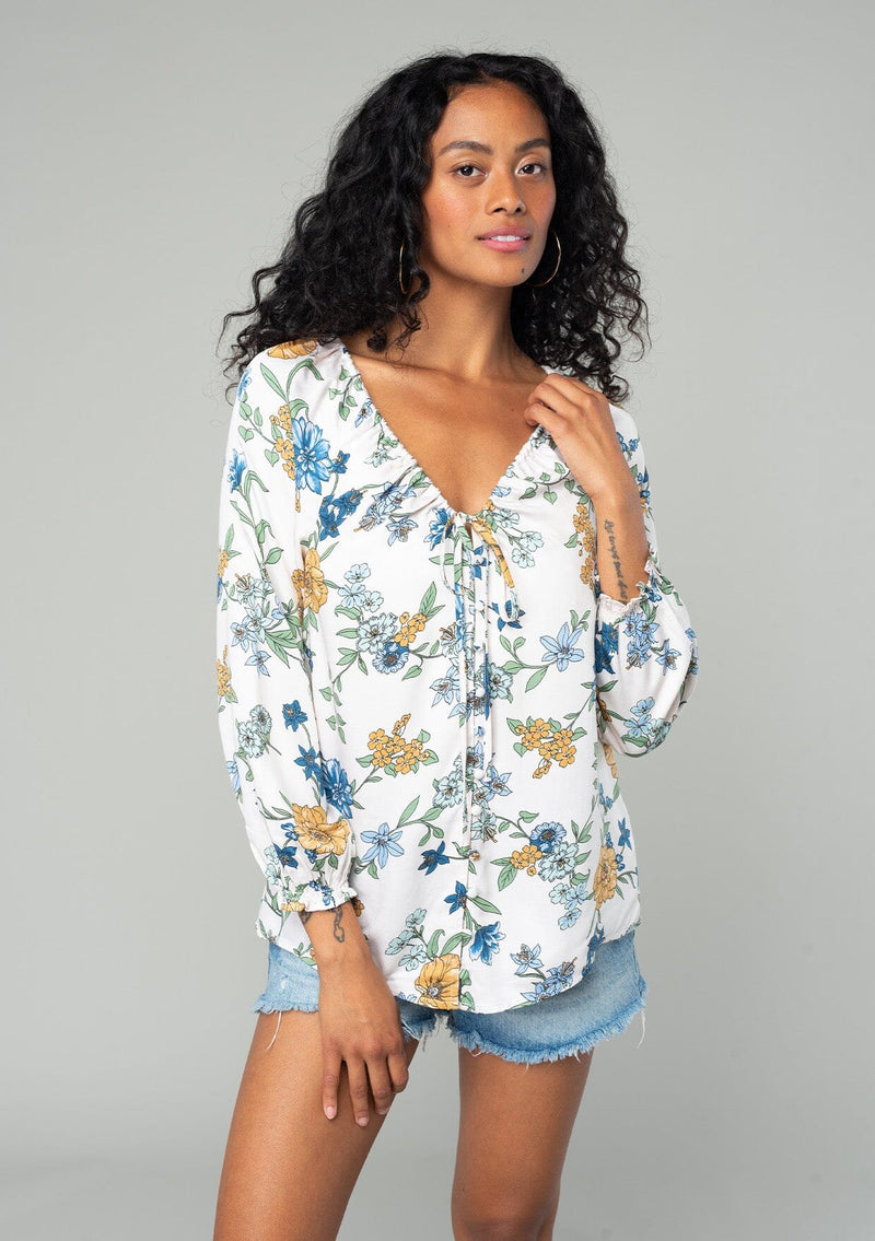 [Color: Natural/Mustard] A half body front facing image of a brunette model wearing a silky bohemian blouse in a natural, blue and yellow floral print. With long sleeves, a ruffled elastic wrist cuff, a self covered button front, and a drawstring v neckline with adjustable tie. 