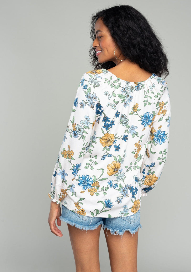 [Color: Natural/Mustard] A back facing image of a brunette model wearing a silky bohemian blouse in a natural, blue and yellow floral print. With long sleeves, a ruffled elastic wrist cuff, a self covered button front, and a drawstring v neckline with adjustable tie. 