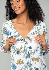 [Color: Natural/Mustard] A close up front facing image of a brunette model wearing a silky bohemian blouse in a natural, blue and yellow floral print. With long sleeves, a ruffled elastic wrist cuff, a self covered button front, and a drawstring v neckline with adjustable tie. 