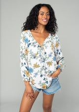 [Color: Natural/Mustard] A front facing image of a brunette model wearing a silky bohemian blouse in a natural, blue and yellow floral print. With long sleeves, a ruffled elastic wrist cuff, a self covered button front, and a drawstring v neckline with adjustable tie. 