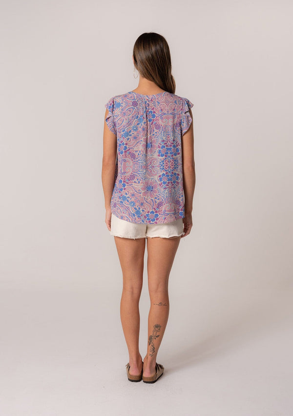 [Color: Dusty Rose/Blue] A back facing image of a brunette model wearing a lightweight summer top in a pink and blue floral print with light catching gold metallic clip dot details. With short ruffle sleeves and a v neckline. 