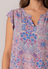 [Color: Dusty Rose/Blue] A close up front facing image of a brunette model wearing a lightweight summer top in a pink and blue floral print with light catching gold metallic clip dot details. With short ruffle sleeves and a v neckline. 
