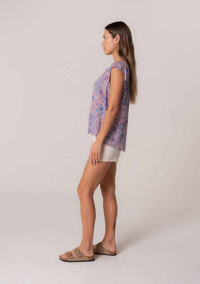 [Color: Dusty Rose/Blue] A side facing image of a brunette model wearing a lightweight summer top in a pink and blue floral print with light catching gold metallic clip dot details. With short ruffle sleeves and a v neckline. 