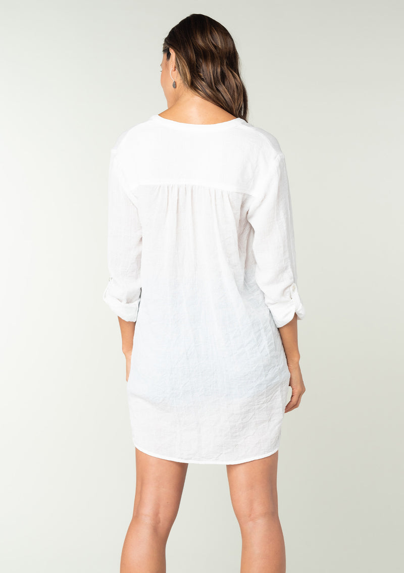 [Color: White] A back facing image of a brunette model wearing a classic lightweight white tunic shirt. With long sleeves, a button tab sleeve, a button front, a v neckline, and side pockets. 