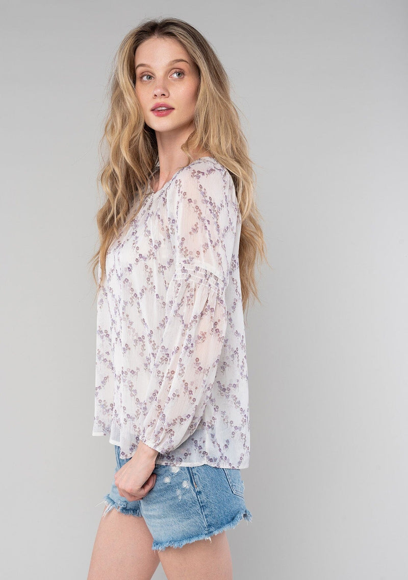 [Color: Ivory/Plum] A side facing image of a blonde model wearing a sheer chiffon spring bohemian blouse in an ivory and plum purple floral print. With long bishop sleeves, a wide smocked elastic neckline, a front keyhole detail with button closure, and a flowy relaxed fit. 