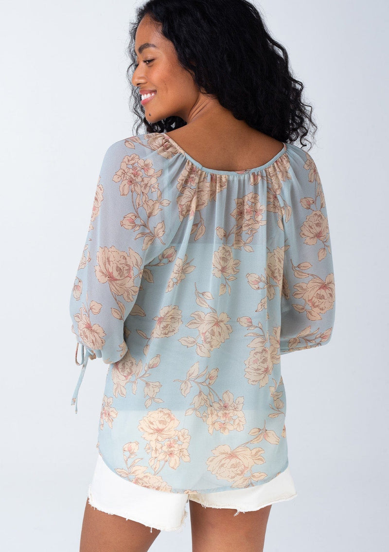 [Color: Dusty Blue/Natural] A back facing image of a brunette model wearing a sheer chiffon spring bohemian blouse in a dusty blue and natural floral print. With three quarter length sleeves, adjustable tie cuffs, a v neckline, and a flowy relaxed fit. 