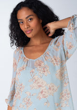 [Color: Dusty Blue/Natural] A close up front facing image of a brunette model wearing a sheer chiffon spring bohemian blouse in a dusty blue and natural floral print. With three quarter length sleeves, adjustable tie cuffs, a v neckline, and a flowy relaxed fit. 