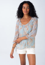 [Color: Dusty Blue/Natural] A front facing image of a brunette model wearing a sheer chiffon spring bohemian blouse in a dusty blue and natural floral print. With three quarter length sleeves, adjustable tie cuffs, a v neckline, and a flowy relaxed fit. 
