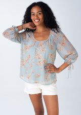 [Color: Dusty Blue/Natural] A half body front facing image of a brunette model wearing a sheer chiffon spring bohemian blouse in a dusty blue and natural floral print. With three quarter length sleeves, adjustable tie cuffs, a v neckline, and a flowy relaxed fit. 