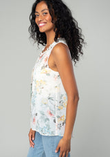 [Color: Natural/Dusty Sage] A side facing image of a brunette model wearing a sleeveless chiffon tank top in a natural and sage green floral print. With a ruffle trimmed neckline, a button front, and a split neckline. 