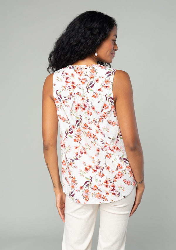 [Color: Natural/Rose] A back facing image of a brunette model wearing a sleeveless blouse in a natural and rose pink floral print. With a self covered button front, a split neckline, and a banded collar. 