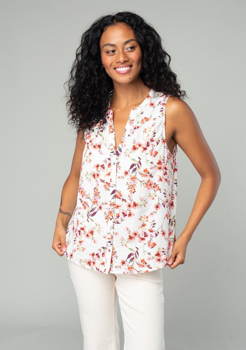 [Color: Natural/Rose] A half body front facing image of a brunette model wearing a sleeveless blouse in a natural and rose pink floral print. With a self covered button front, a split neckline, and a banded collar. 