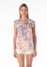 [Color: Natural/Rust] A front facing image of a blonde model wearing a lightweight cotton spring top in a pink and blue floral print. With short raglan sleeves, a ruffled neckline, lace trim, and a v neckline with double tie detail. 