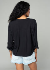 [Color: Black] A back facing image of a brunette model wearing a basic black blouse with long rolled sleeves, a button tab closure, a tie waist detail, and a concealed button front.  