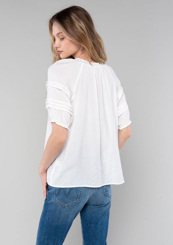 [Color: White] A back facing image of a blonde model wearing a classic bohemian white spring top with short puff sleeves, a button front, a split neckline with tassel ties, and delicate pintuck details. 