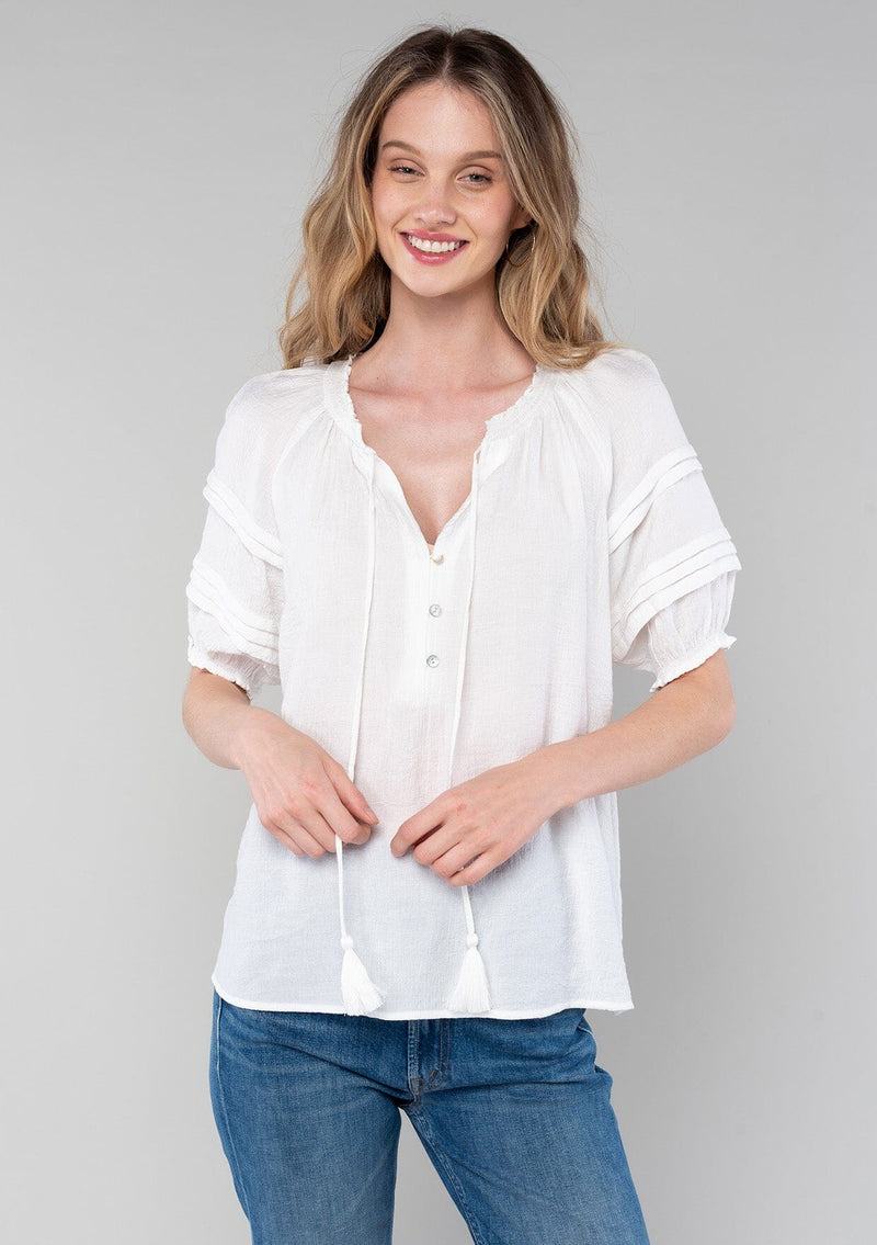[Color: White] A half body front facing image of a blonde model wearing a classic bohemian white spring top with short puff sleeves, a button front, a split neckline with tassel ties, and delicate pintuck details. 