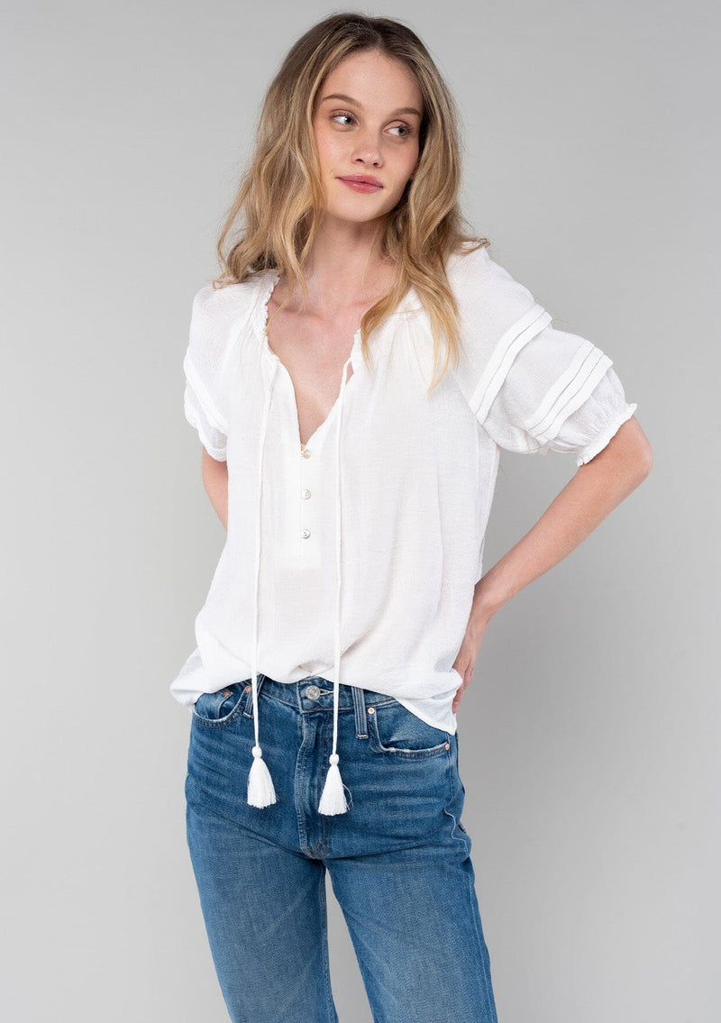 [Color: White] A front facing image of a blonde model wearing a classic bohemian white spring top with short puff sleeves, a button front, a split neckline with tassel ties, and delicate pintuck details. 