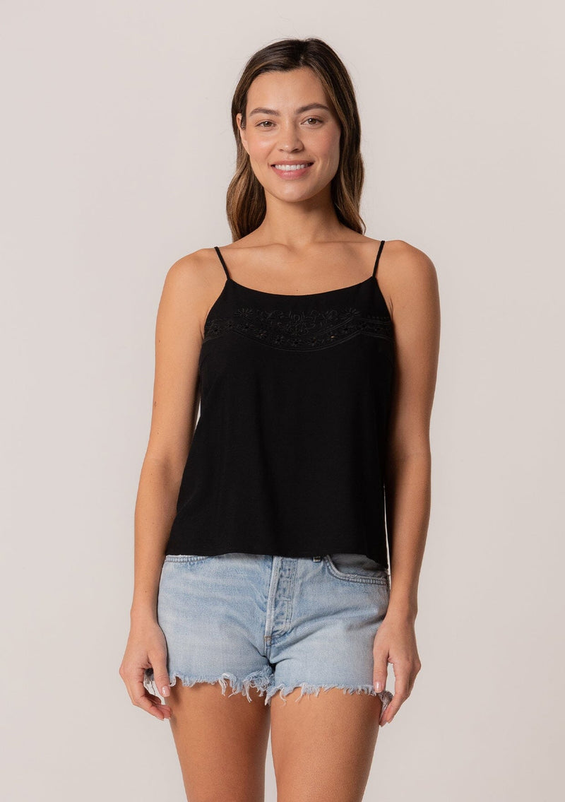 [Color: Black] A front facing image of a brunette model wearing a black bohemian spring camisole tank top with embroidered detail, adjustable spaghetti straps, a scooped neckline, and a self covered button up back detail. The subtle floral embroidery on this cute black boho tank top elevates the style while keeping it simple. The vintage inspired button up back detail will have heads turning this spring/summer season.