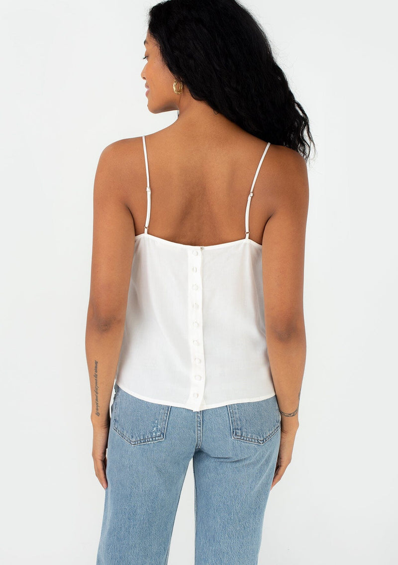 [Color: Off White] A back facing image of a brunette model wearing a bohemian spring camisole tank top with embroidered detail, adjustable spaghetti straps, a scooped neckline, and a self covered button up back detail. 