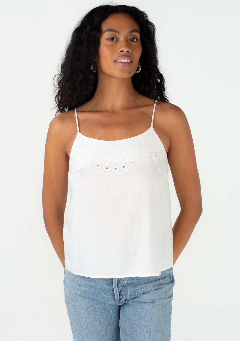 [Color: Off White] A front facing image of a brunette model wearing a bohemian spring camisole tank top with embroidered detail, adjustable spaghetti straps, a scooped neckline, and a self covered button up back detail.  If you're searching for a simple yet unique white spring tank top, you're found it! The subtle flora embroidered neckline elevates any outfit, and the button back details give it a vintage inspired twist.