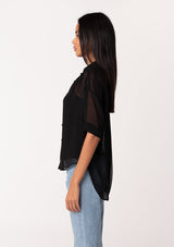 [Color: Black] A side facing image of a brunette model wearing a black sheer chiffon short sleeve button front blouse. With a self covered button front and a collared neckline.