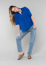 [Color: Cobalt] A full body front facing image of a blonde model wearing a bright blue sheer chiffon short sleeve button front blouse. With a self covered button front and a collared neckline.