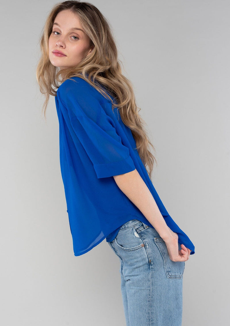 [Color: Cobalt] A side facing image of a blonde model wearing a bright blue sheer chiffon short sleeve button front blouse. With a self covered button front and a collared neckline.