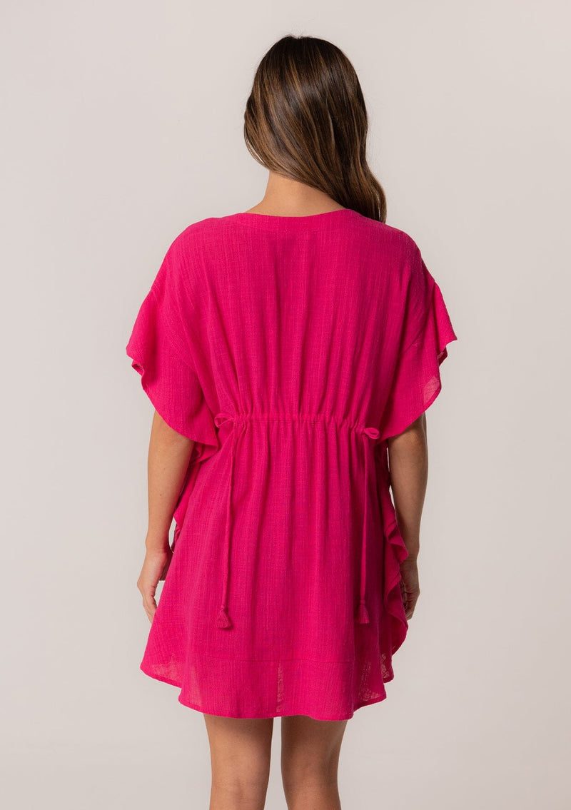 [Color: Fuchsia] A back facing image of a brunette model wearing a bohemian caftan top in a bright pink cotton. A beach cover up style with short sleeves, a ruffled hemline, a v neckline, embroidered details throughout, and a drawstring waist detail in the front and back with tassel ties.