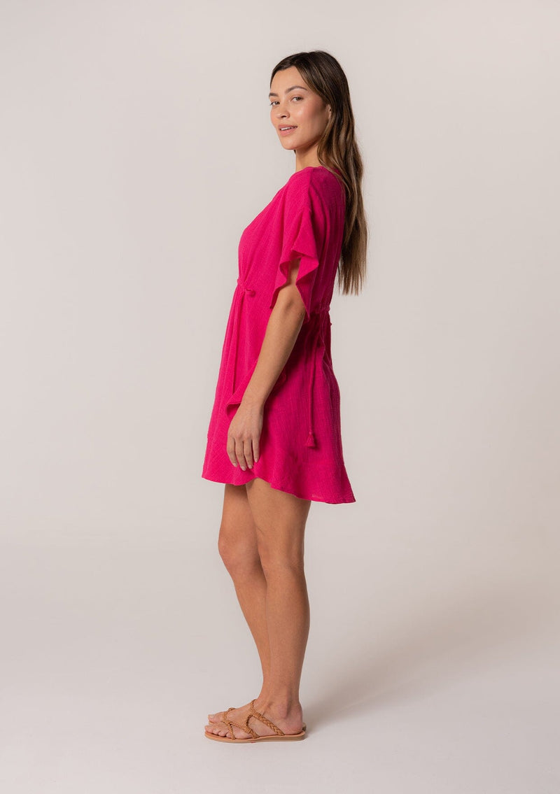 [Color: Fuchsia] A full body side facing image of a brunette model wearing a bohemian caftan top in a bright pink cotton. A beach cover up style with short sleeves, a ruffled hemline, a v neckline, embroidered details throughout, and a drawstring waist detail in the front and back with tassel ties.