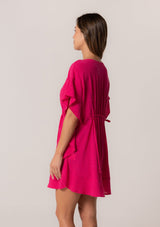 [Color: Fuchsia] A side facing image of a brunette model wearing a bohemian caftan top in a bright pink cotton. A beach cover up style with short sleeves, a ruffled hemline, a v neckline, embroidered details throughout, and a drawstring waist detail in the front and back with tassel ties.