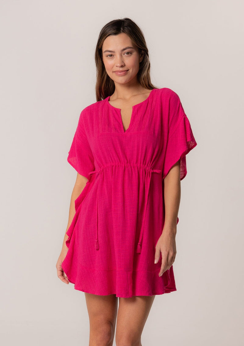 [Color: Fuchsia] A front facing image of a brunette model wearing a bohemian caftan top in a bright pink cotton. A beach cover up style with short sleeves, a ruffled hemline, a v neckline, embroidered details throughout, and a drawstring waist detail in the front and back with tassel ties.