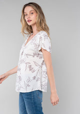 [Color: Natural/Taupe] A side facing image of a blonde model wearing a flowy bohemian spring top in a natural and taupe floral print. With short drop sleeves, a round neckline, and a self covered button front. 