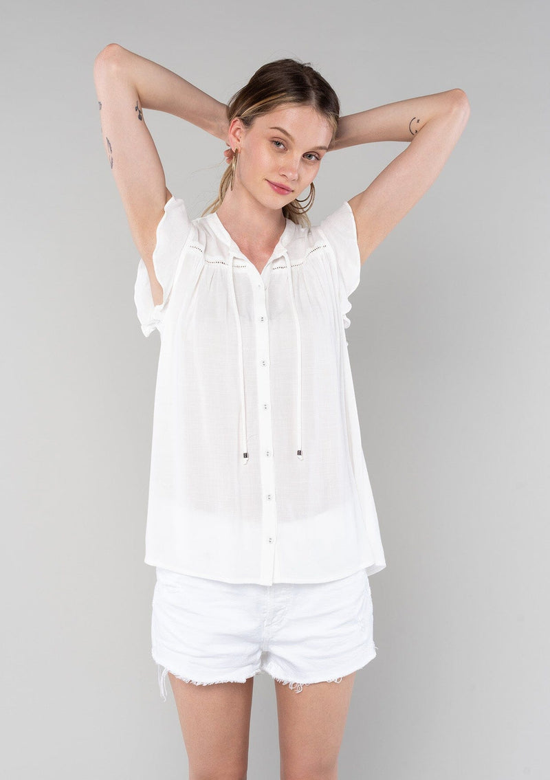 [Color: Vanilla] A front facing image of a blonde model wearing a solid white bohemian spring top. With short flutter cap sleeves, a self covered button front, neckline ties, and a relaxed fit. 