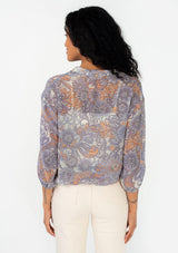 [Color: Ivory/Dusty Plum] A back facing image of a brunette model wearing a sheer chiffon bohemian blouse in a retro inspired purple floral print. With three quarter length sleeves, a collared neckline, a button front, and a drawstring waist with adjustable tie. 