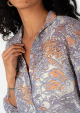 [Color: Ivory/Dusty Plum] A close up side facing image of a brunette model wearing a sheer chiffon bohemian blouse in a retro inspired purple floral print. With three quarter length sleeves, a collared neckline, a button front, and a drawstring waist with adjustable tie. 