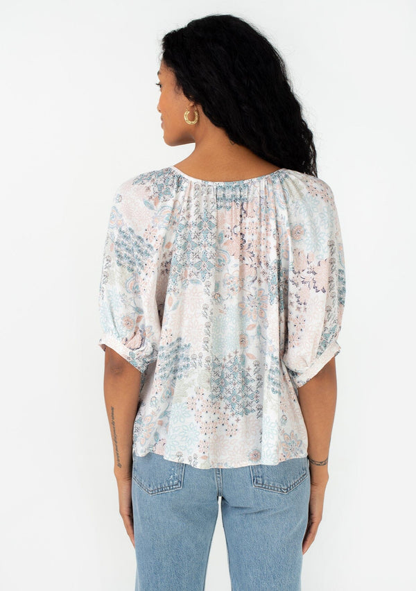 [Color: Dusty Blue/Teal] A back facing image of a brunette model wearing a bohemian spring blouse in a blue floral print. With half length puff sleeves, a button front, a round neckline, and a flowy relaxed fit. 