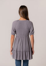 [Color: Light Grey] A back facing image of a brunette model wearing a bohemian spring tunic top in a light grey crinkled fabric. With short puff sleeves, ruffle trim, a v neckline, a smocked bodice, an empire waist, and a flowy tiered body.