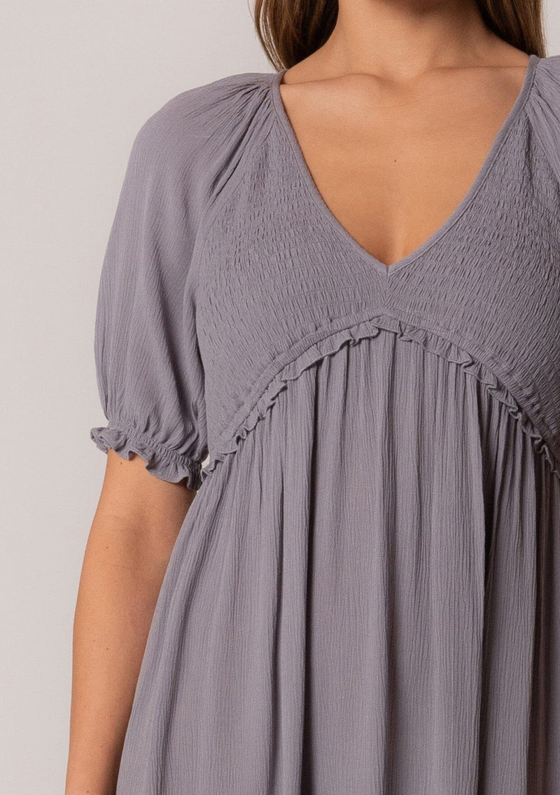 [Color: Light Grey] A close up front facing image of a brunette model wearing a bohemian spring tunic top in a light grey crinkled fabric. With short puff sleeves, ruffle trim, a v neckline, a smocked bodice, an empire waist, and a flowy tiered body.