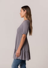 [Color: Light Grey] A side facing image of a brunette model wearing a bohemian spring tunic top in a light grey crinkled fabric. With short puff sleeves, ruffle trim, a v neckline, a smocked bodice, an empire waist, and a flowy tiered body.
