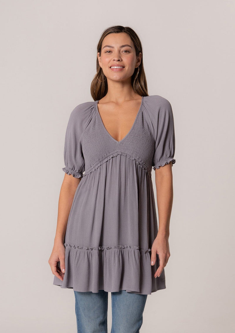 [Color: Light Grey] A front facing image of a brunette model wearing a bohemian spring tunic top in a light grey crinkled fabric. With short puff sleeves, ruffle trim, a v neckline, a smocked bodice, an empire waist, and a flowy tiered body.