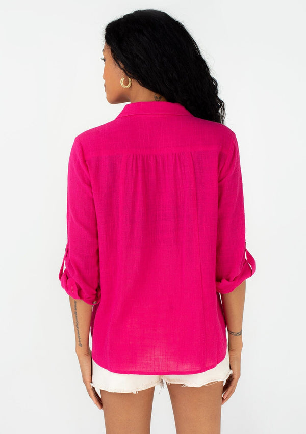 [Color: Fuchsia] A back facing image of a brunette model wearing a bright pink cotton shirt. With long rolled sleeved, a button tab sleeve closure, a button front, two front patch pockets, a tie front waist detail, and a high low hemline. 