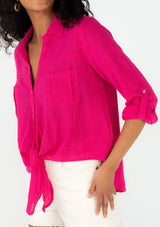 [Color: Fuchsia] A close up front facing image of a brunette model wearing a bright pink cotton shirt. With long rolled sleeved, a button tab sleeve closure, a button front, two front patch pockets, a tie front waist detail, and a high low hemline. 