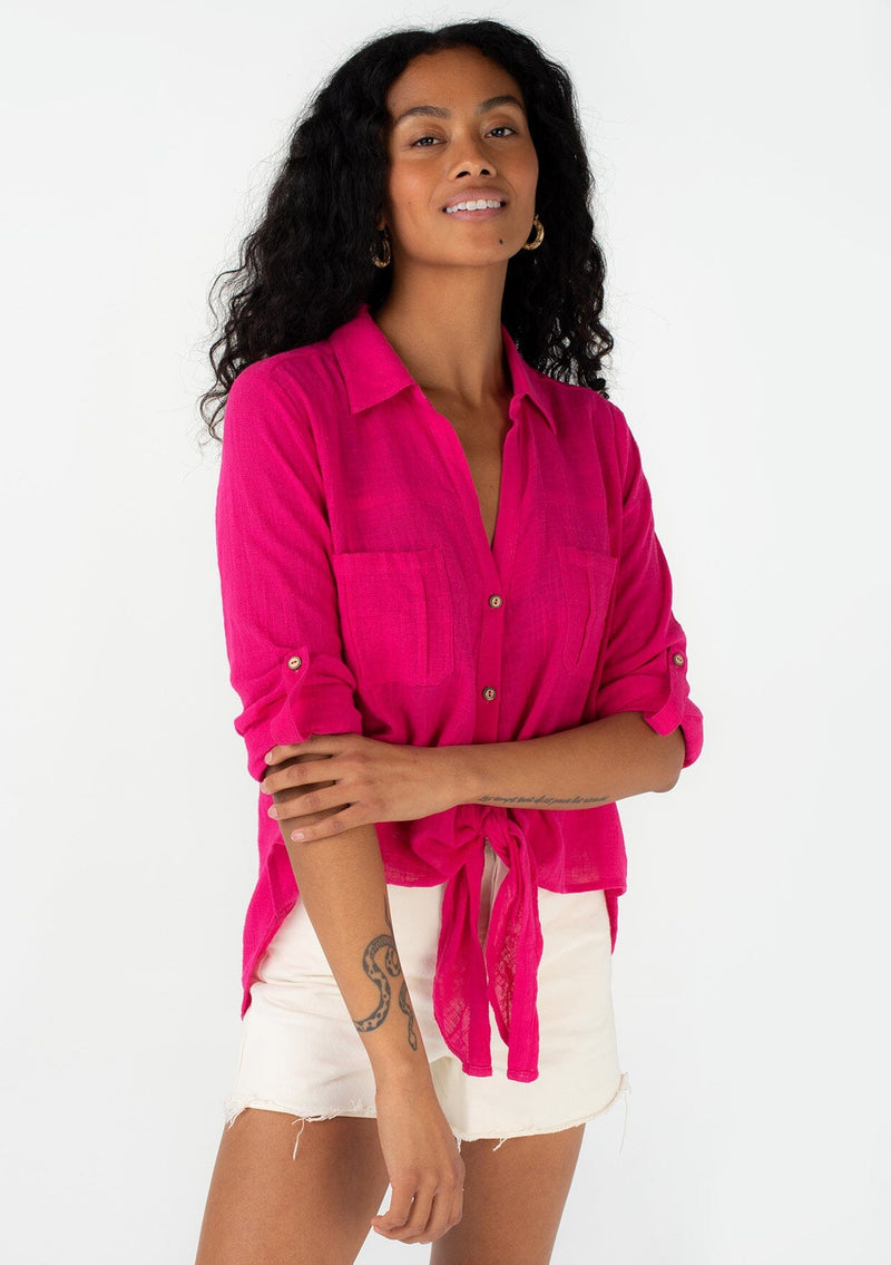 [Color: Fuchsia] A front facing image of a brunette model wearing a bright pink cotton shirt. With long rolled sleeved, a button tab sleeve closure, a button front, two front patch pockets, a tie front waist detail, and a high low hemline. 