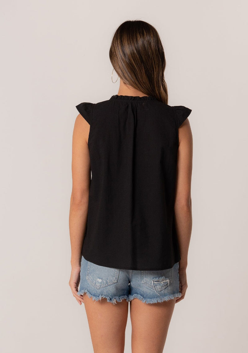 [Color: Black] A back facing image of a brunette model wearing a bohemian cotton spring top in a black colorway. With short cap sleeves, a self covered button front, a ruffled neckline, a v neckline, and embroidered detail.